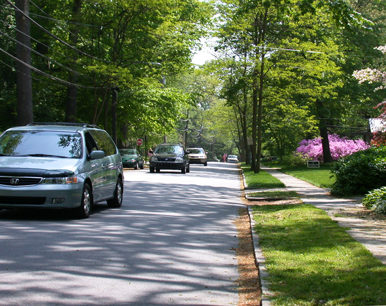 A suburban street scene with cars receding into the distance, trees lining the road, the road narrowing toward a vanishing point.