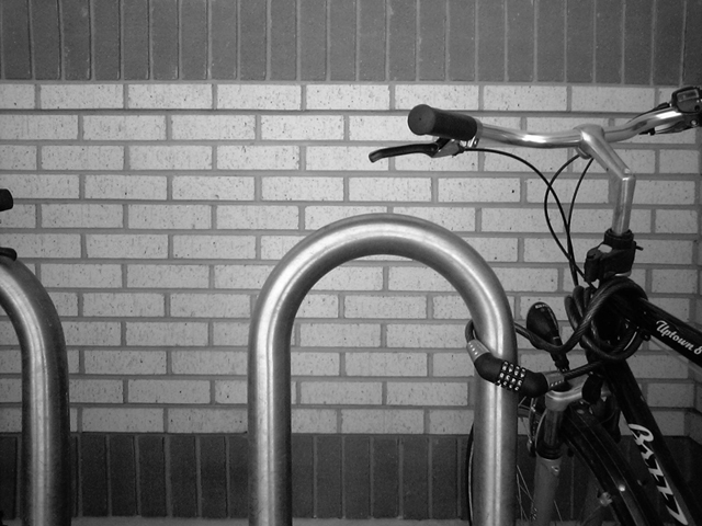 Original grayscale photograph of a bicycle parked in a steel rack against a brick wall.